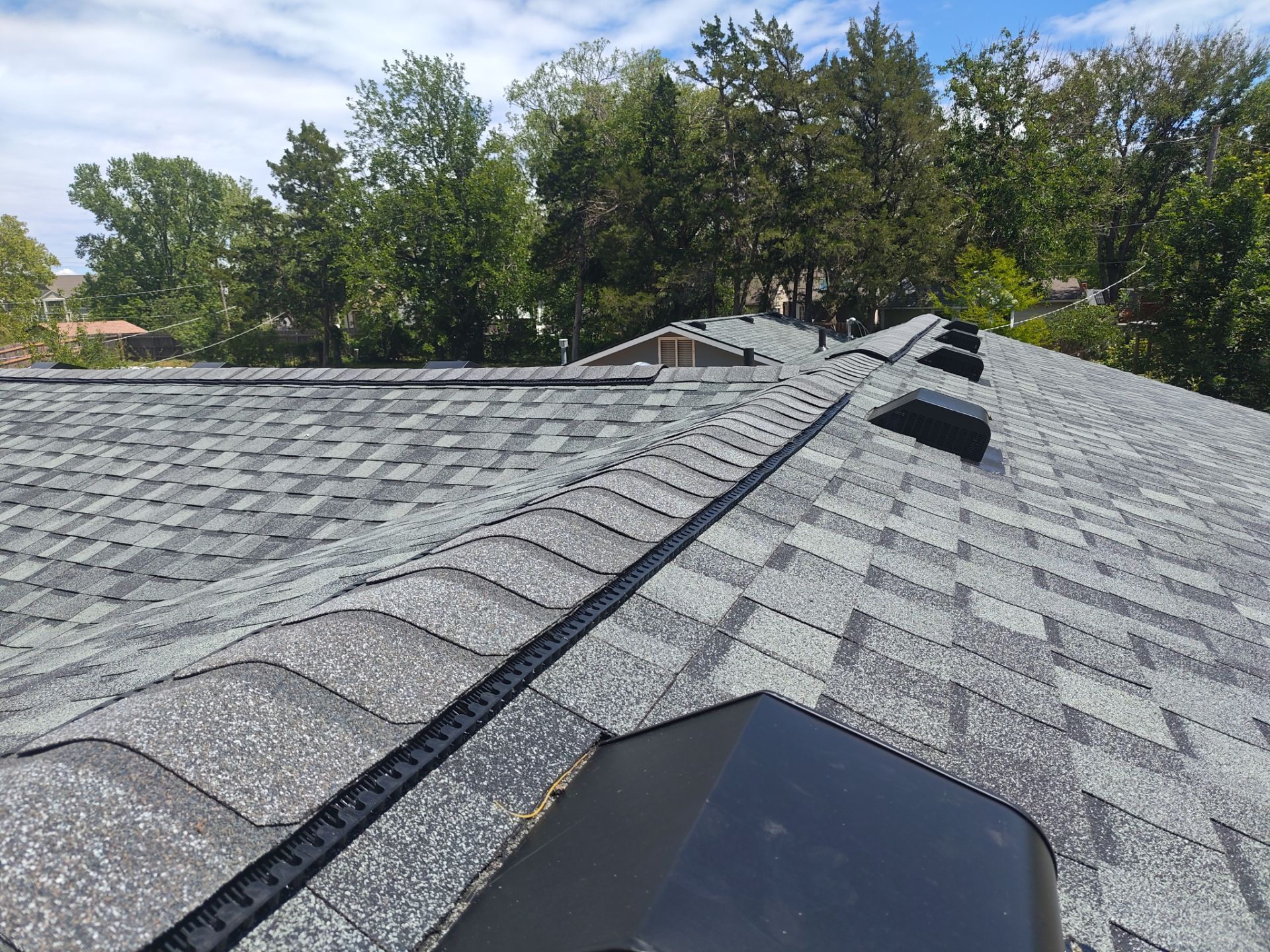 Overhead view of a gray asphalt shingle roof with multiple vents and a curved ridge line, with trees and blue sky in the background.