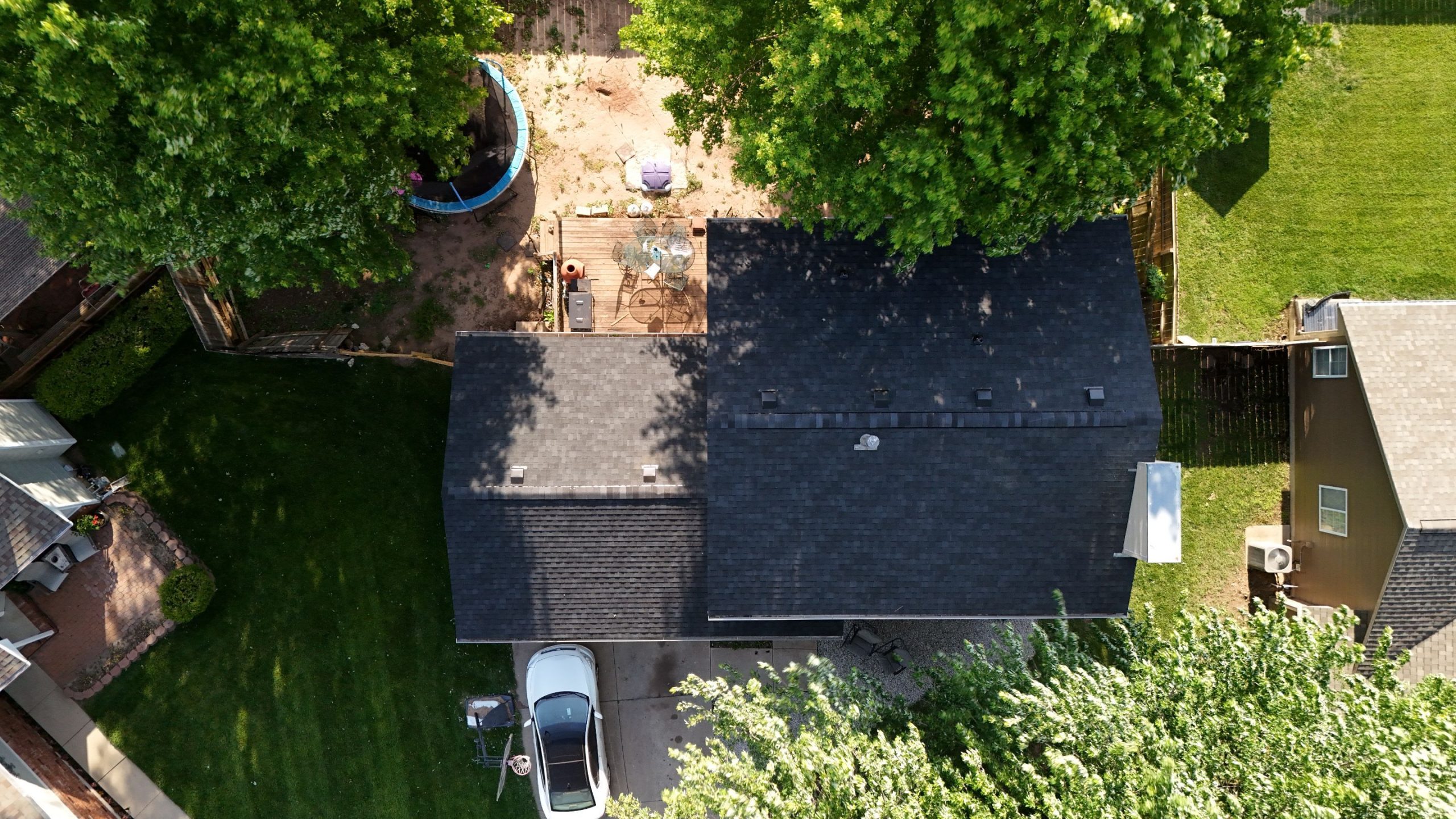 default Solid Rock Commercial Roofing Aerial view of a suburban backyard showing a dark-roofed house, wooden deck with patio furniture, a circular trampoline, parked cars, and green lawns framed by trees.