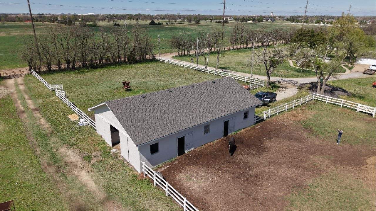 A light-gray barn with a dark roof, surrounded by white fences in a rural field; a person stands near the dirt yard.