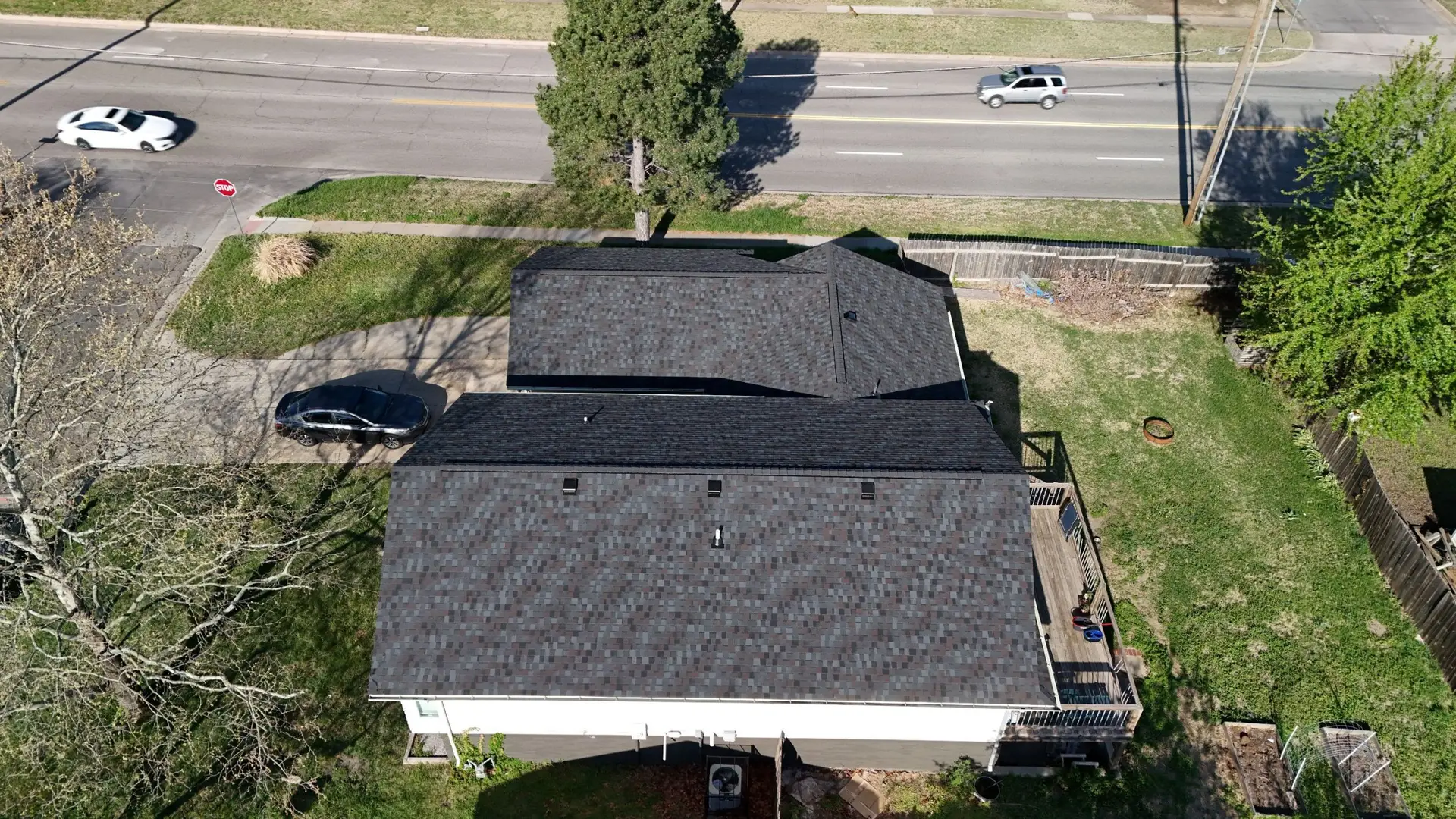 Aerial view of a suburban street: a white car on the road, a dark SUV in a driveway, stop sign at a sidewalk, and a large house with a shaded roof.